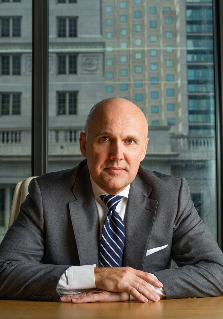 A businessman in a suit sitting at a desk in an office with city buildings outside.